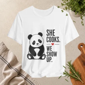 White T-shirt featuring a cute black-and-white panda and the phrase “She Cooks. We Show Up.” with a small red heart.