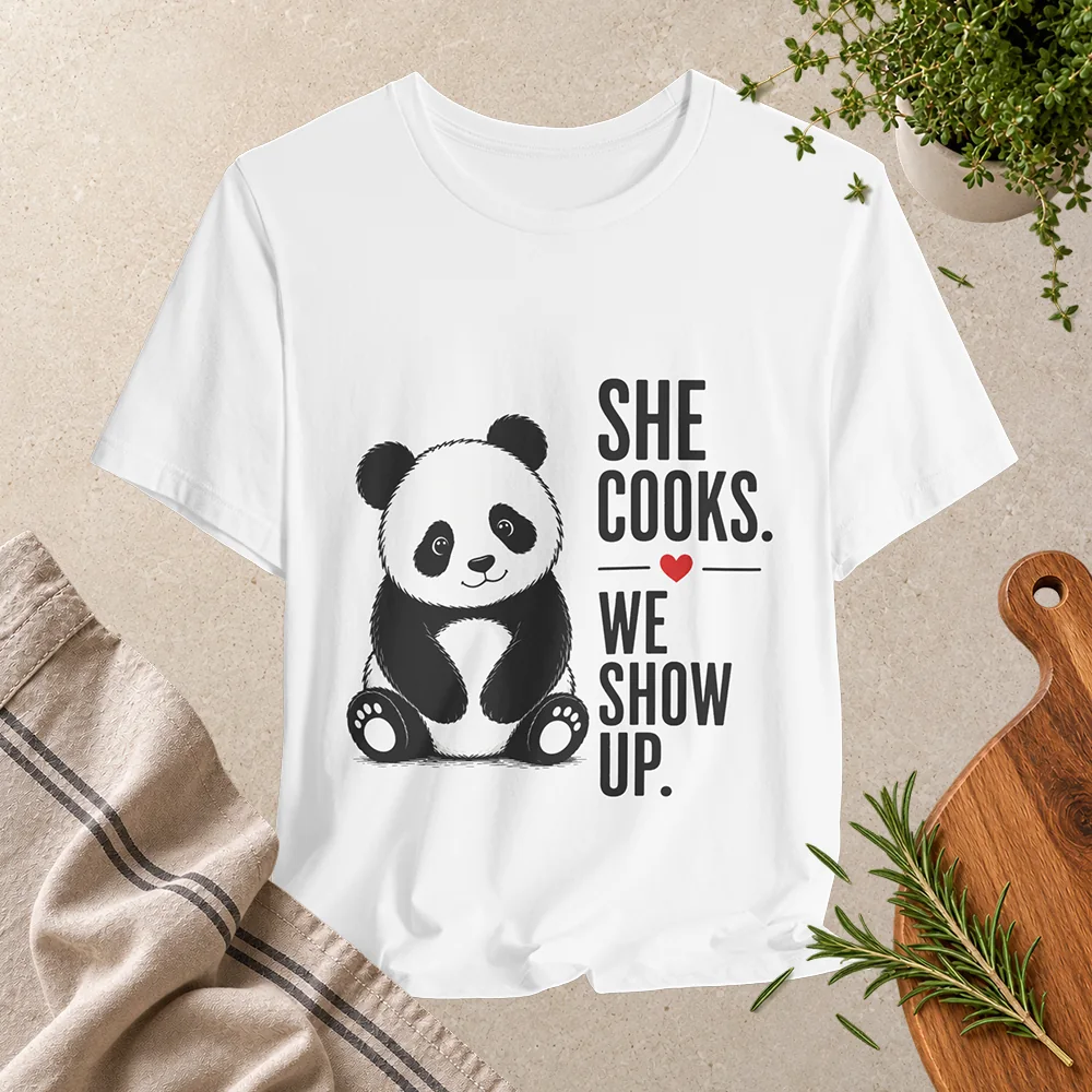 White T-shirt featuring a cute black-and-white panda and the phrase “She Cooks. We Show Up.” with a small red heart.
