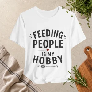 White T-shirt with “Feeding People Is My Hobby” typography design, featuring a small red heart and spoon graphic, displayed in a kitchen-inspired flat lay setting.